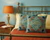 cushions featuring a coloured lino-cut design of four rainbow bee-eaters facing towards the centre of the cushion surrounded by kapok flowers and buds on a blue background on a brass bed with an orange bedspread
