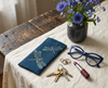 Canvas glasses slip case featuring pale blue stylised dragonflies on a dark blue patterned background on a cloth with a pair of blue glasses, a set of keys, some rings, a lipstick and a bunch of blue flowers in a dark coloured vase