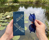 Canvas glasses slip case featuring pale blue stylised dragonflies on a dark blue patterned background and a pair of dark blue glasses behind held by hands in front of a billabong with lilies
