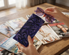 Canvas glasses slip case featuring a leafy pattern of deep sea kelp with bubbles in purples held in a woman's hand against a backdrop of magazines scatted on a table