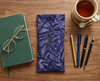 Canvas glasses slip case featuring a leafy pattern of deep sea kelp with bubbles in purples on a table with a notepad, pens and a cup of tea