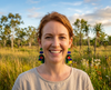 A smiling woman wearing acrylic earrings featuring stylised Gouldian finches