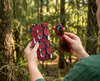 Canvas glasses slip case covered in red mushrooms with white spots on a dark teal green background being held up with a pair of red glasses in a pine forest
