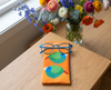 Canvas glasses slip case featuring very round and highly stylised blue-faced parrotfinches on an orange background on a table with a pair of blue glasses. Behind are two glass vases with colourful flowers in them.