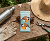 Canvas glasses case featuring two highly stylised and very round black throated finches on a pale blue background on a rustic timber bench with sunglasses, secateurs, gardening gloves, a water bottle and a battered sunhat