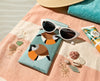 Canvas glasses case featuring two highly stylised and very round black throated finches on a pale blue background on a beach towel with white sunglasses, shells and sunblock