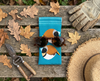 Canvas glasses slip case featuring two very round stylised chestnut breasted mannikins on a blue background on a bench with a pair of secateurs, a pair of gardening gloves, a straw hat and a pair of brown sunglasses