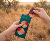 Canvas glasses slip case featuring two very round and highly stylised female crimson finches on a teal green background - a pair of red sunglasses are being removed from the pouch against an natural Australian grassland background