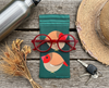 Canvas glasses slip case featuring two very round and highly stylised female crimson finches on a teal green background on a wooden bench with a pair of red spectacles, car keys, a battered sun hat, a water bottle and a small bunch of dried Australian native flowers