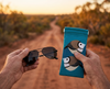 Canvas glasses slip case featuring two very round and highly stylised double barred finches on a blue background, held in a man's hand alongside a pair of spectacles against an out of focus Australian bush background with a dirt track.