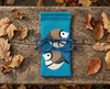 Canvas glasses slip case featuring two very round and highly stylised double barred finches on a blue background on a piece of timber surrounded by dried leaves. A pair of blue spectacles sits on top of the glasses case