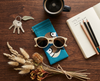 Canvas glasses slip case featuring two very round and highly stylised double barred finches on a blue background on a bench with a notebook, sunglasses, a cup of coffee, a bunch of keys, a notebook and pencils and a bunch of dried Australian flowers and seedpods