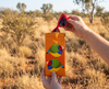 Canvas glasses slip case featuring two very round stylised gouldian finches with red faces on an orange background being held by one hand as a pair of red glasses are removed from them. The background is a savanna grassland.