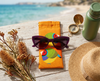 Canvas glasses slip case featuring two very round stylised gouldian finches with red faces on an orange background on a bench or table near the sea with a pair of purple sunglasses, a sunhat, a compass, a water bottle and a bunch of dried grasses and flowers