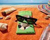Canvas glasses slip case featuring two very round and highly stylised long-tailed finches on a green background on an orange beach towel with black sunglasses, a hat, sunblock and some shells