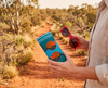 Canvas glasses slip case featuring two very round highly stylised painted firetail finches on a blue background, being held along with a pair of red glasses on a dirt track in the Australian bush