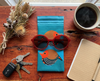 Canvas glasses slip case featuring two very round highly stylised painted firetail finches on a blue background on red timber bench with a mug of coffee, a small bunch of dried native Australian flowers, a note book, a pair of red glasses and a set of keys