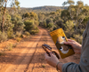 Canvas glasses slip case featuring two very round highly stylised plum-headed finches on a mustard background being held by a woman with a pair of tortoiseshell glasses in her left hand against an Australian bush scene with a dirt track