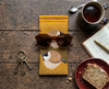 Canvas glasses slip case featuring two very round highly stylised plum-headed finches on a mustard background on a rustic bench with a pair of brown glasses, a mug of tea, a notebook and pen, a piece of cake on a plate and a small bunch of old keys