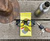 Canvas glasses slip case featuring two very round highly stylised red-browed finches on a yellow background on a rough timber bench with an old leather hat, a drink bottle, a pair of black rimmed glasses and a set of car keys