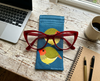 Canvas glasses slip case featuring two very round and highly stylised star finches on a blue background on a bench with a mug of coffee, a notebook with pens, a pair of red glasses and a laptop