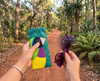 Canvas glasses slip case featuring a large Gouldian finch with a red face against a teal background of triangles held in a woman's hand. The other hand holds a pair of purple sunglasses. In the background is a dirt track winding into tropical bushland.
