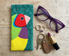 Canvas glasses slip case featuring a large Gouldian finch with a red face against a teal background of triangles on a timber surface with keys, rings, lipstick and a pair of glasses