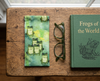 Canvas glasses slip case featuring several green tree frogs against a green watery background with some small clumps of grass, on a bench with a pair of green glasses and a book titled 'Frogs of the World'