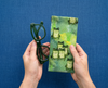 Canvas glasses slip case featuring several green tree frogs against a green watery background with some small clumps of grass, held in a hand alongside a pair of green spectacles on a blue background