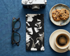 A canvas glasses slip case covered with a black and white magpie goose design on a blue cloth with a pair of black spectacles, a camera, a saucer with home baked biscuits and a black cup of coffee