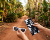 Canvas glasses slip case covered with a black and white magpie goose design held by a woman who also holds white sunglasses.  A dirt road leads into a tropical scene in the background