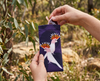 A pair of pink sunglasses being removed from a canvas glasses slip case featuring two stylised pink cockatoos with their combs up against a purple background. In the background is an Australian bush scene.