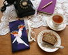 A canvas glasses slip case featuring two stylised pink cockatoos with their combs up against a purple background lies on a table with a pair of white glasses, a cup of tea and slice of cake, an old fashioned phone and a purple notebook with a gold pen.