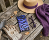 A canvas glasses slip case showing rows of tall purple mushrooms against a black background on a timber bench with a pair of gardening gloves, a hat, scarf, trowel and a pair of purple spectacles