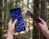 A canvas glasses slip case showing rows of tall purple mushrooms against a black background held in a hand. A second hand holds a pair of purple sunglasses. In the background is a pine forest.