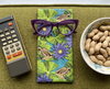 Canvas glasses slip case featuring a pattern of striped rocket frogs on waterlily pads with purple waterlily  flowers on a green couch with a pair of purple glasses, a remote control and a bowl of peanuts