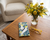 Canvas glasses slip case featuring two sulphur crested cockatoos perched in Red Box tree on a coffee table with a pair of yellos glasses and a vase of yellow native Australian flowers