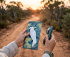 Canvas glasses slip case featuring a sulphur crested cockatoo perched in Red Box tree, held in a woman's hand. The other hand holds a pair of sunglasses. In the background is a dirt track leading through the Australian bush towards a sunset.