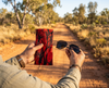 A man's hands hold a pair of sunglasses and a canvas glasses slip case with a black and red pattern of pandanus trees covering it. In the background a dirt road stretches off into the Australian bush