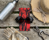A canvas glasses slip case with a black and red pattern of pandanus trees covering it lies on a bench beneath a pair of black, round sunglasses. They are surrounded by a hat, a pair of gardening gloves, a pair of secateurs and a water bottle.