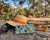 A canvas glasses slip case featuring coloured lino prints of two rainbow bee-eaters surrounded by kapok flowers on a log with a pair of orange sunglasses, a straw hat with an orange ribbon and a pair of binoculars, in the Australian bush