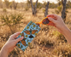 A woman's hand pulls a pair of orange sunglasses from a canvas glasses slip case featuring coloured lino prints of two rainbow bee-eaters surrounded by kapok flowers. In the background is an Australian bush scene.