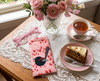 A canvas glasses slip case featuring a stylised red-backed fairywren against a pink pattern of speargrass seed heads sits on a lace tablecloth alongside a pair of pink spectacles, a cup of tea, piece of cake on a small plate and a bunch of pink flowers