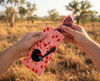 A pair of pink sunglasses being pulled from a canvas glasses slip case featuring a stylised red-backed fairywren against a pink pattern of speargrass seed heads. The background is a natural Australia grassland scene.