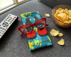 Canvas glasses slip case featuring a stylised pair of red-tailed black cockatoos perched in a wattle on  teal green/blue background on a couch with a remote control, a pair of red glasses and a bowl of chips