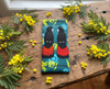 Canvas glasses slip case featuring a stylised pair of red-tailed black cockatoos perched in a wattle on  teal green/blue background on a timber bench with wattle flowers