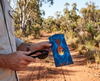 A man holding a pair of black sunglasses and a canvas glasses slip case featuring a stylised silver-crowned friarbird on a blue patterned background. A dirt road in the Australian bush stretches into the distance behind him