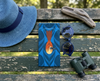 A canvas glasses slip case featuring a stylised silver-crowned friarbird on a blue patterned background lies on a painted park bench with a hat, a pair of blue sunglasses, a pair of binoculars and a piece of driftwood