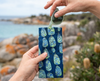 A pair of pale green sunglasses being pulled from a canvas glasses case featuring a semi abstract design based on bubbleweed in blues and greens. In the background is an out-of-focus beach scene with grey and orange rocks