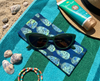 Canvas glasses case featuring a semi abstract design based on bubbleweed in blues and greens on a striped beachtowel with a pair of dark green glasses, a sunhat, sunblock, shells and beads